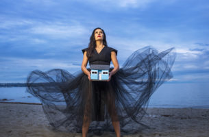 Quelemia Sparrow in a black dress holding a small blue model of a house