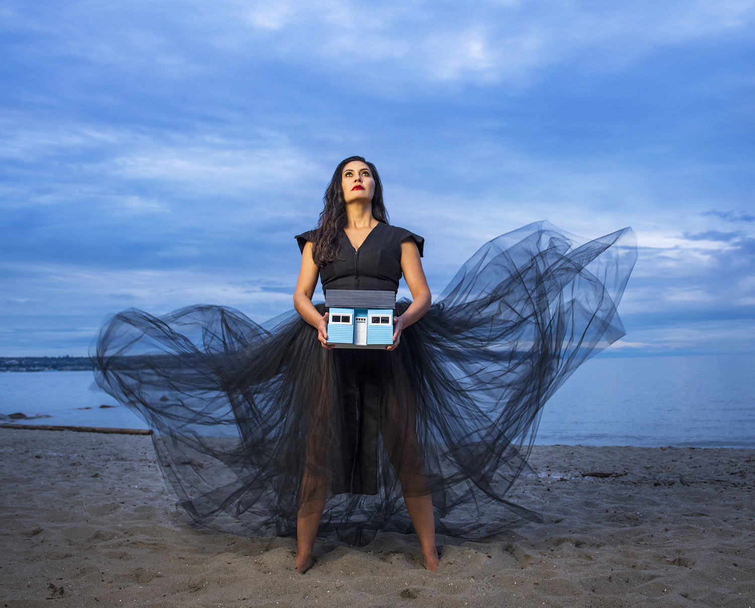 Quelemia Sparrow in a black dress holding a small blue model of a house