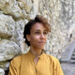 A woman of colour, wearing a yellow blouse stands against a stone wall, looking off camera