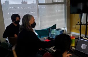 Two teenagers working with an adult mentor on laptops, using a design application