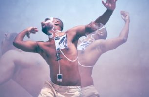 A layered photo of a brown man with black and white squares painted on his body. He is captured dancing in three poses. The foreground shows him gracefully swaying his arms upward. On the left, his upper body is curved towards the sky. On the right, he is looking up his face showing white paint, with his arm raised in a fist.