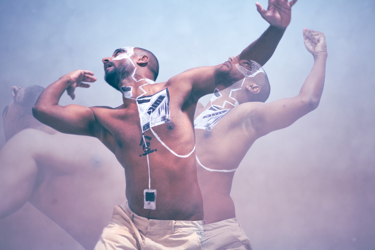 A layered photo of a brown man with black and white squares painted on his body. He is captured dancing in three poses. The foreground shows him gracefully swaying his arms upward. On the left, his upper body is curved towards the sky. On the right, he is looking up his face showing white paint, with his arm raised in a fist.