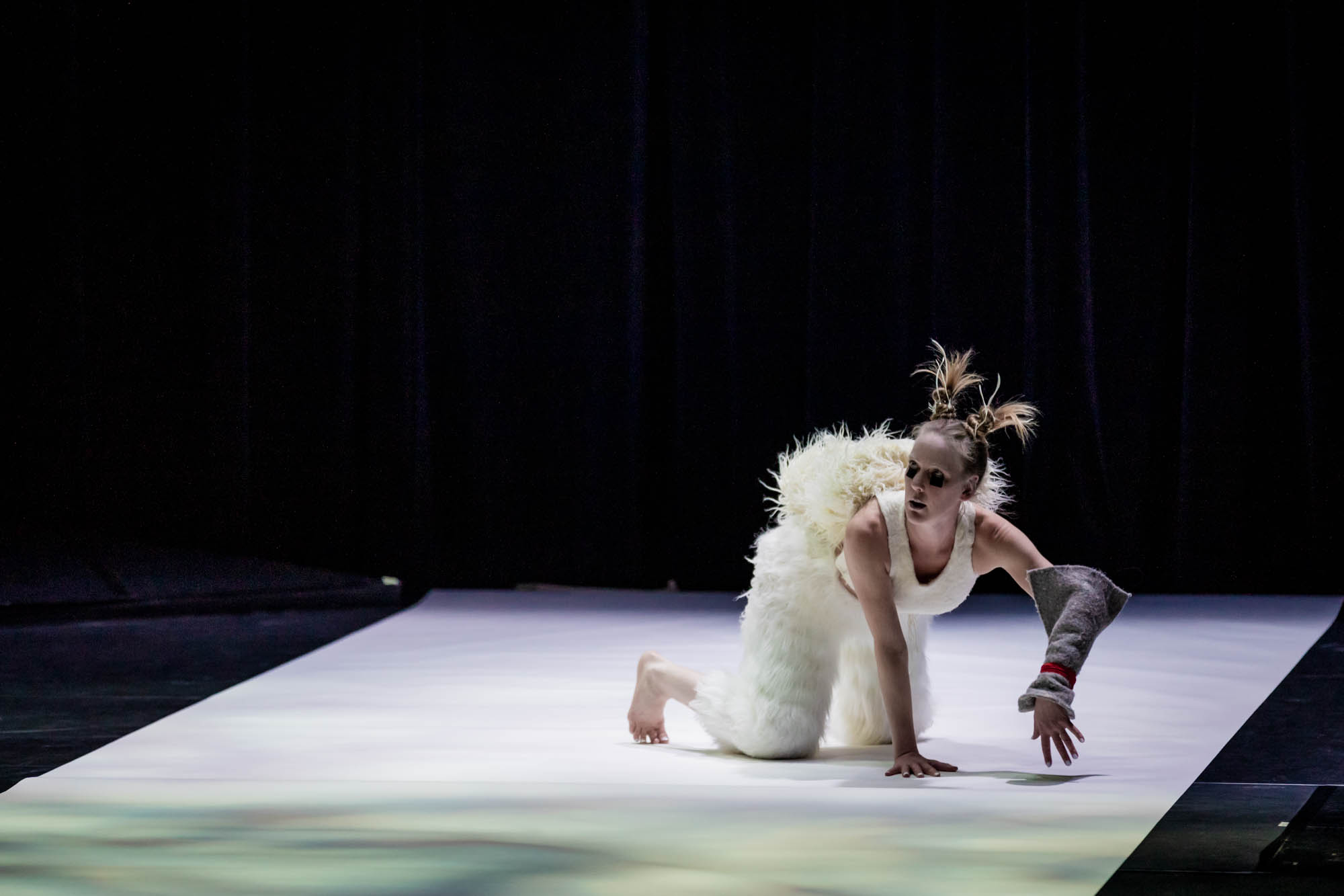 A performer with blonde pigtails wears dark eye makeup, white tank top, furry white paints, and a grey knitted sleeve on the left arm. They are kneeling on the ground looking up. The ground is lit with white lights amid a dark stage.