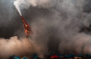 A figure wearing a salmon-coloured shirt and face mask holds up a red tube exhuming a large cloud of white smoke that envelops the room. On the ground are multicoloured pieces of crinkled plastic.