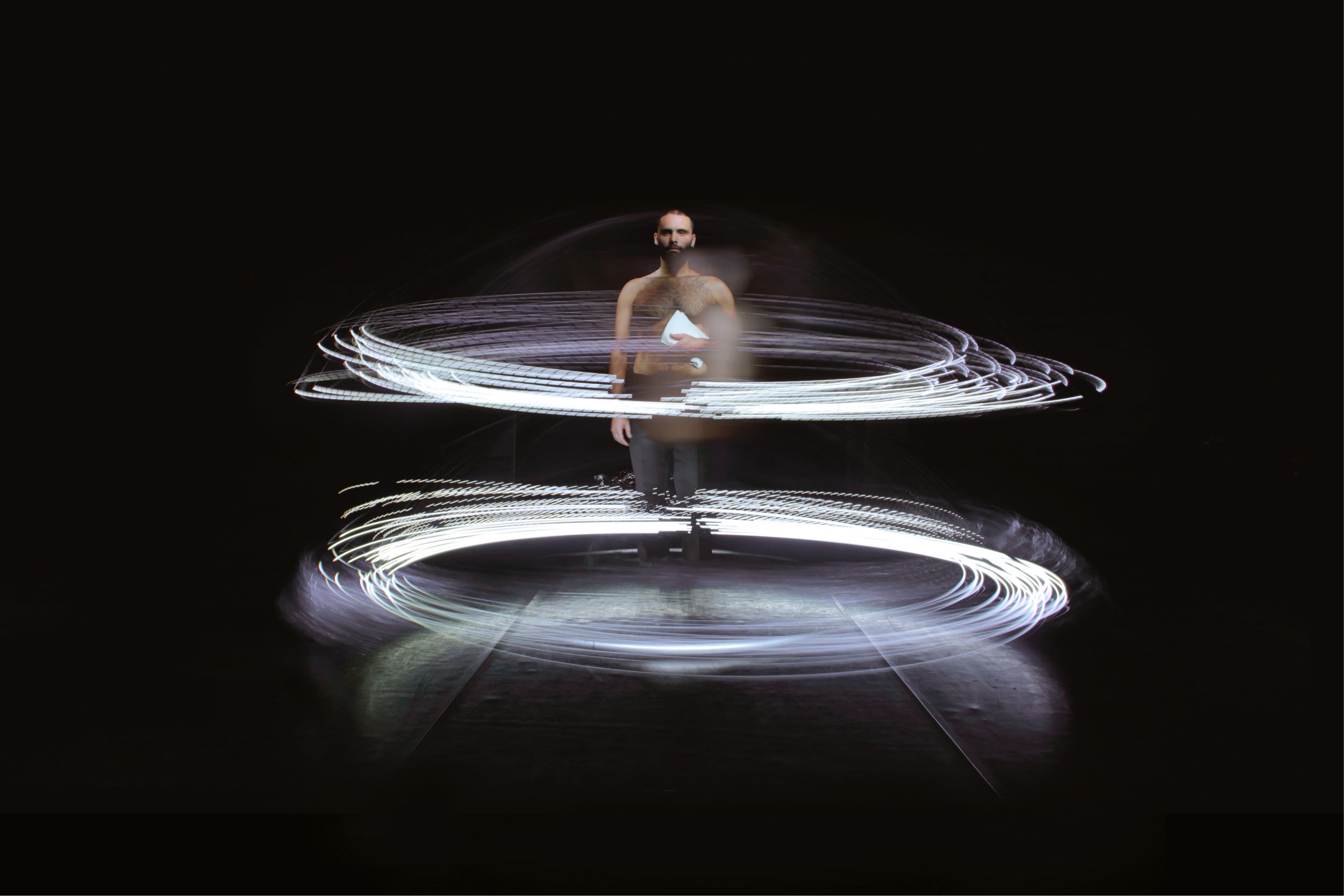 Long-exposure image of Juan Ignacio Tula in Instante, standing in a dark room. He is shirtless and stares into the camera, holding a white stack of paper. Circling him are two rings made of many horizontal rings of light motion lines.