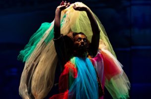 A dancer wearing a costume of brightly coloured flowing tulle stands with arms raised, looking up. They are lit by light above amid a dark blue background.