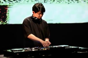 Jaha Koo, who has short black hair and is wearing glasses and a black T-shirt and headphones around his beck, is DJing. In the background is a video of green glitches.