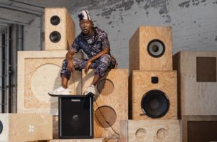 nora chipaumire, wearing a white and black-patterned jumpsuit, thick black necklaces, white and black head covering, and white sneakers, looks into the camera. she sits on top of a high stack of wood stereo speakers, feet resting on top of a black speaker.