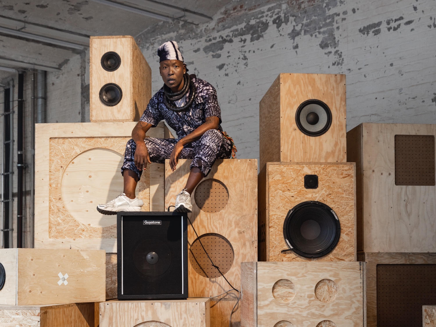 nora chipaumire, wearing a white and black-patterned jumpsuit, thick black necklaces, white and black head covering, and white sneakers, looks into the camera. she sits on top of a high stack of wood stereo speakers, feet resting on top of a black speaker.