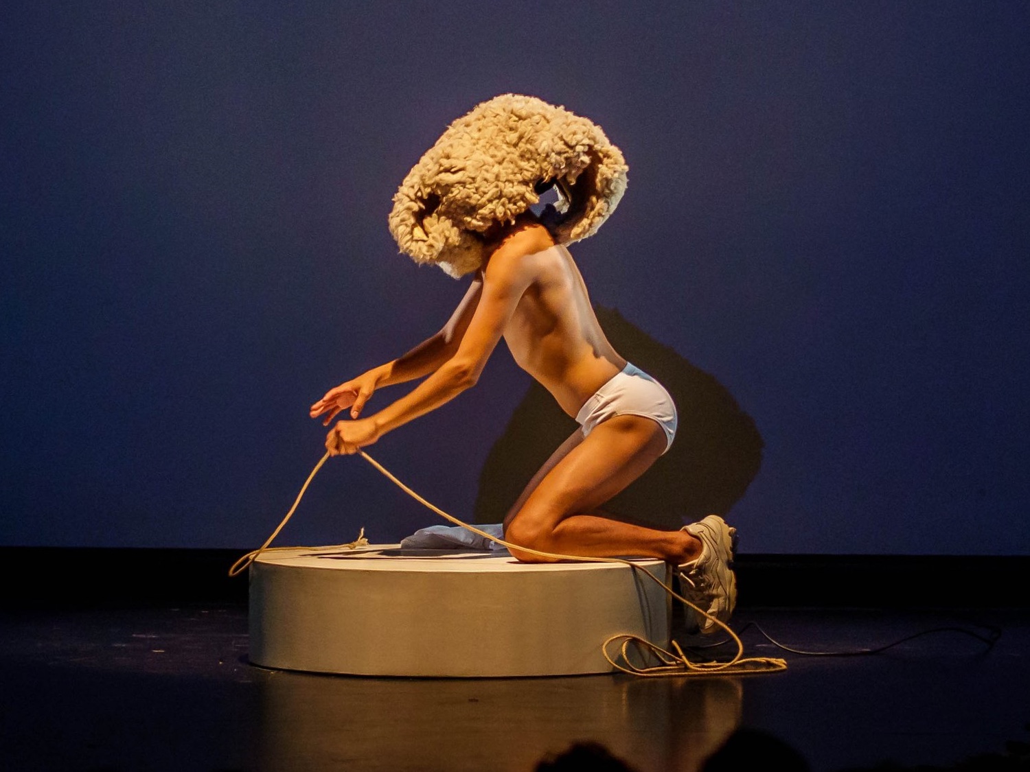 Performer Tiziano Cruz, wearing white briefs and white sneakers, kneels on a small round platform. He pulls on a piece of rope and is wearing a large woolen piece on his head, obscuring the face.