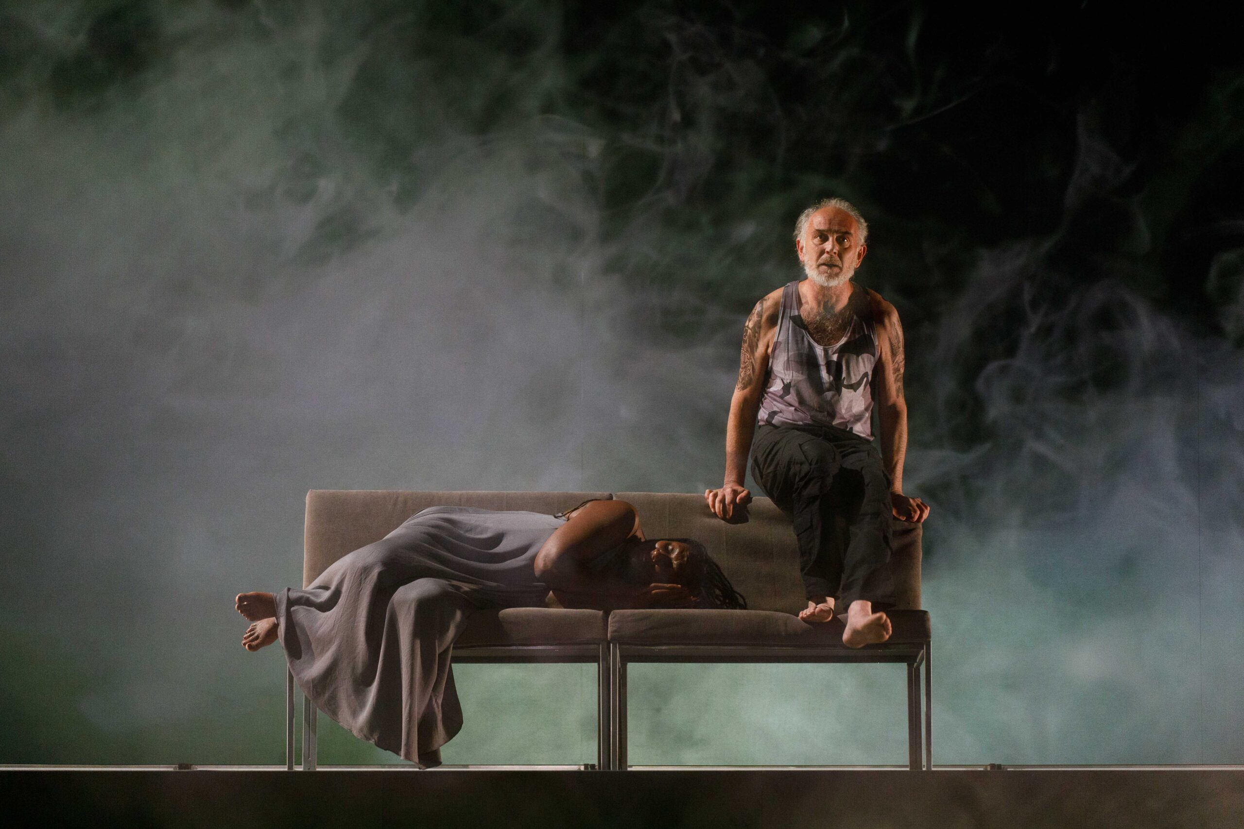 A man with graying hair sits on the backrest of the sofa clutching it tightly. Beside him, is a Black woman who appears to be sleeping, her feet hanging off the edge of the seat.