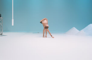 A wide shot of a person contorting their body. They are bending down to reach for the floor, with their torso tilted to the right.