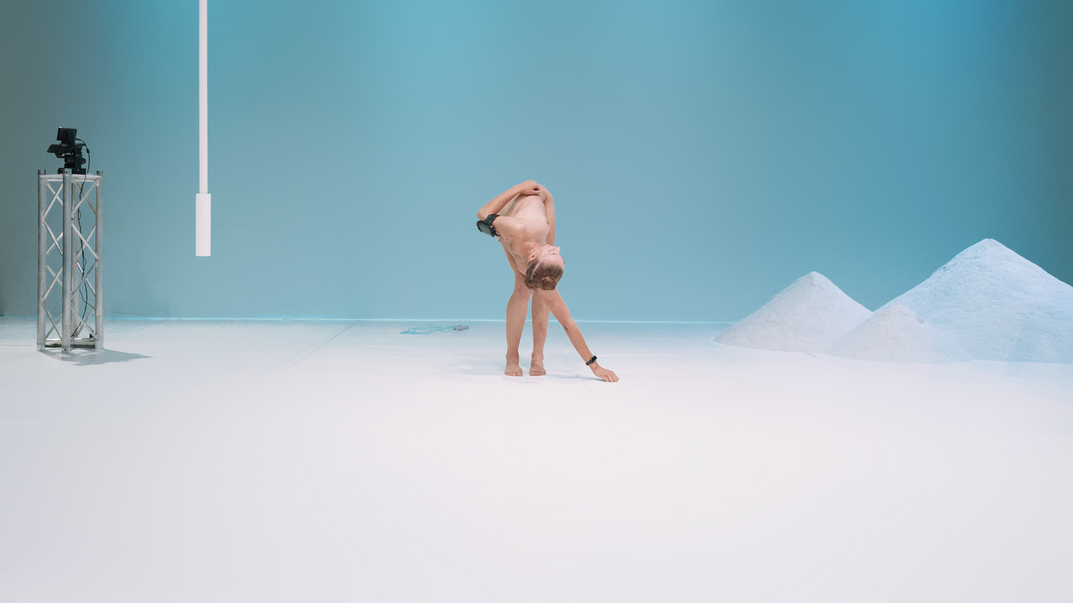 A wide shot of a person contorting their body. They are bending down to reach for the floor, with their torso tilted to the right.