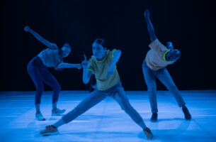 A wide shot of three performers looking directly into the camera in various poses. The performer in the centre is a woman looking directly into the camera. She is in a wide stance, holding up one finger on each hand. The performer on the left is twisting her body towards the center. The performer on the right is a man lunging forward with both his arms raised behind his back.