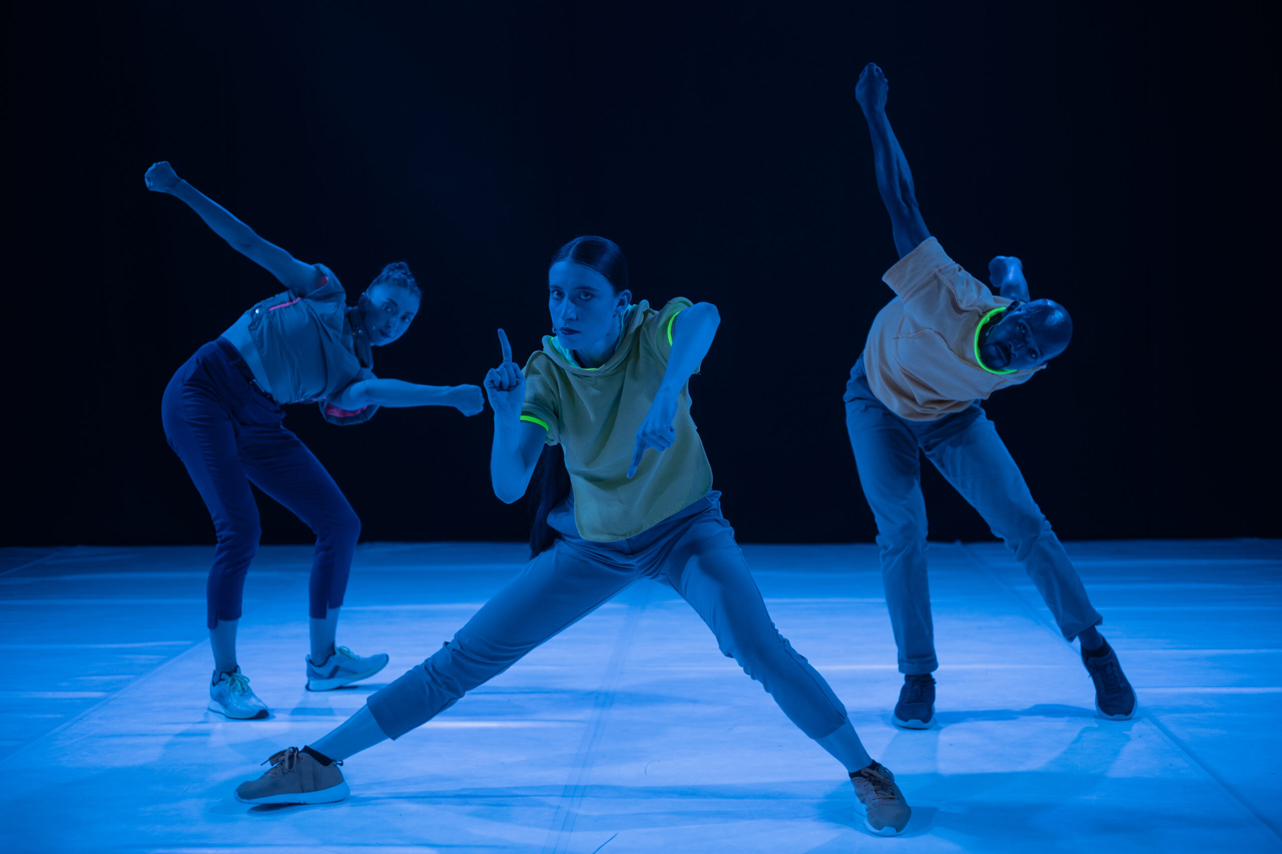 A wide shot of three performers looking directly into the camera in various poses. The performer in the centre is a woman looking directly into the camera. She is in a wide stance, holding up one finger on each hand. The performer on the left is twisting her body towards the center. The performer on the right is a man lunging forward with both his arms raised behind his back.