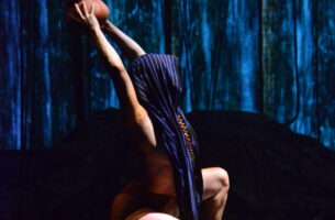 A photo of a woman balancing on two pots. A scarf is draped over her head. She is holding another pot up with stretched arms.