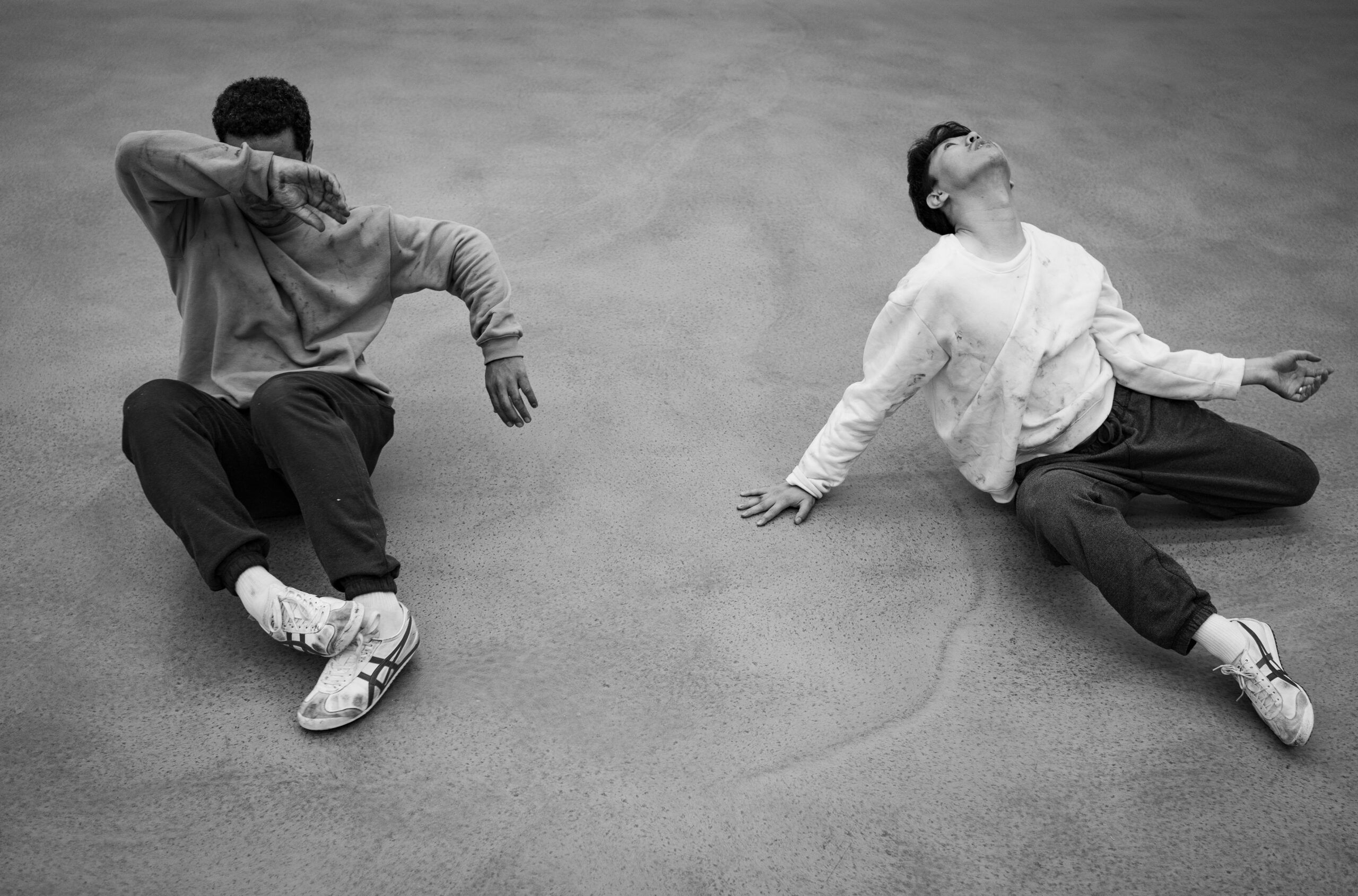 A black and white photo of two performers on the floor. The performer on the left hides their face with their body tensed. The performer on the right is looking up. Their neck is outstretched, with one leg extended, and one hand pressed on the floor.