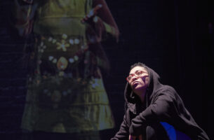 A person half-kneeling with crossed arms while holding a mic. The person is looking up at the light, There is a woman projected behind them.