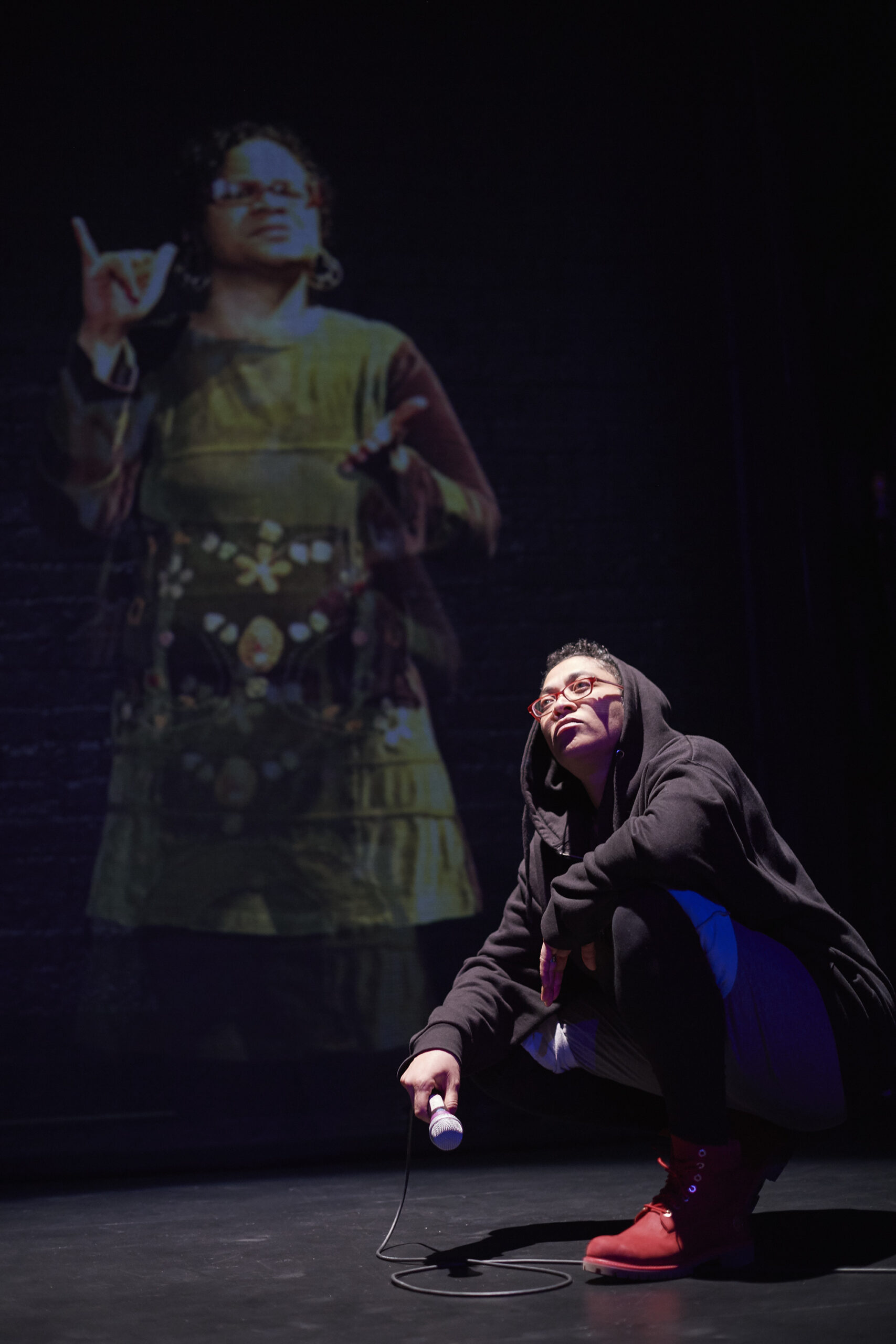 A person half-kneeling with crossed arms while holding a mic. The person is looking up at the light, There is a woman projected behind them.