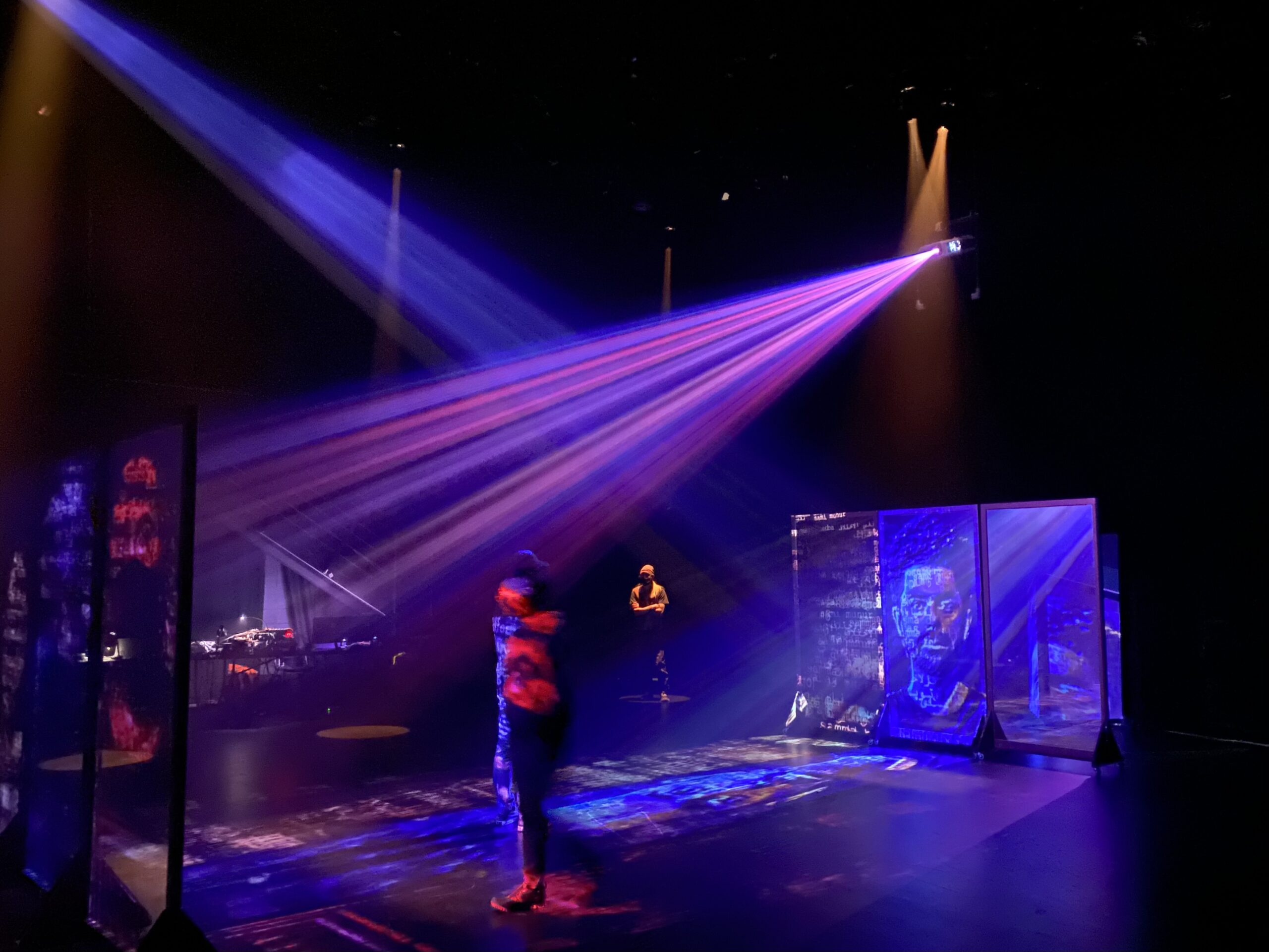 A wide shot of a stage bathed in purple and pink lights. A man’s face is projected onto screens facing each other.