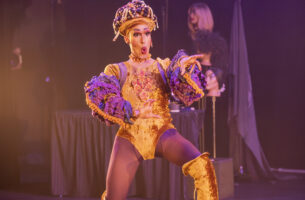 A south-asian drag artist wears a gold, crushed-velvet leotard with matching knee-high high-heeled boots. The artist is caught mid gesture and mid word, reaching out with a look of shock on their face. In the background, an array of wigs perch atop a small cluster of mannequin heads.