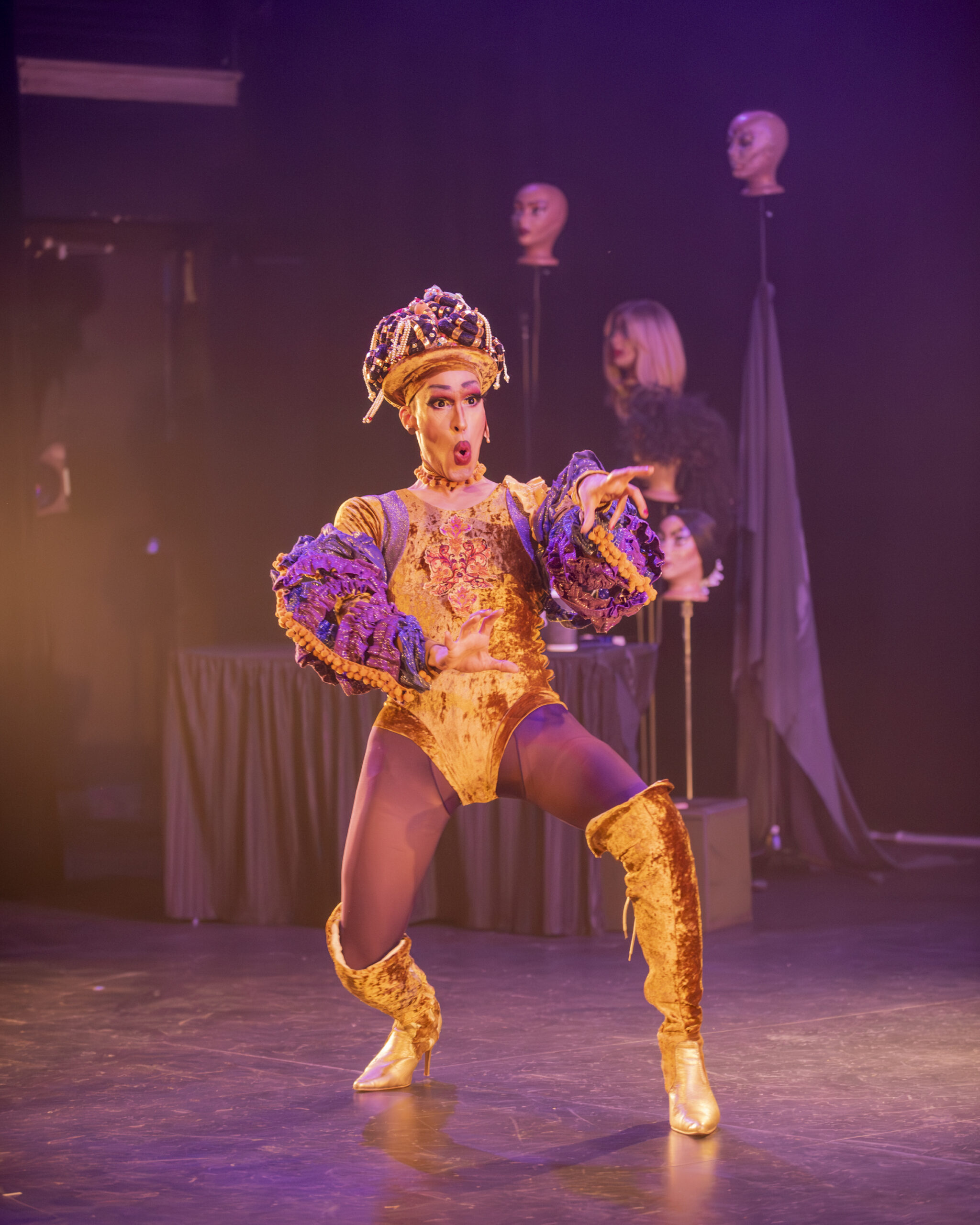 A south-asian drag artist wears a gold, crushed-velvet leotard with matching knee-high high-heeled boots. The artist is caught mid gesture and mid word, reaching out with a look of shock on their face. In the background, an array of wigs perch atop a small cluster of mannequin heads.