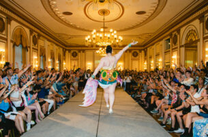 In an opulent ballroom, a performer struts down a runway lined with rows of cheering, grinning audience. Dressed in a colourful short skirt with a hand-painted fish tail, her bare back is visible as she strides away.