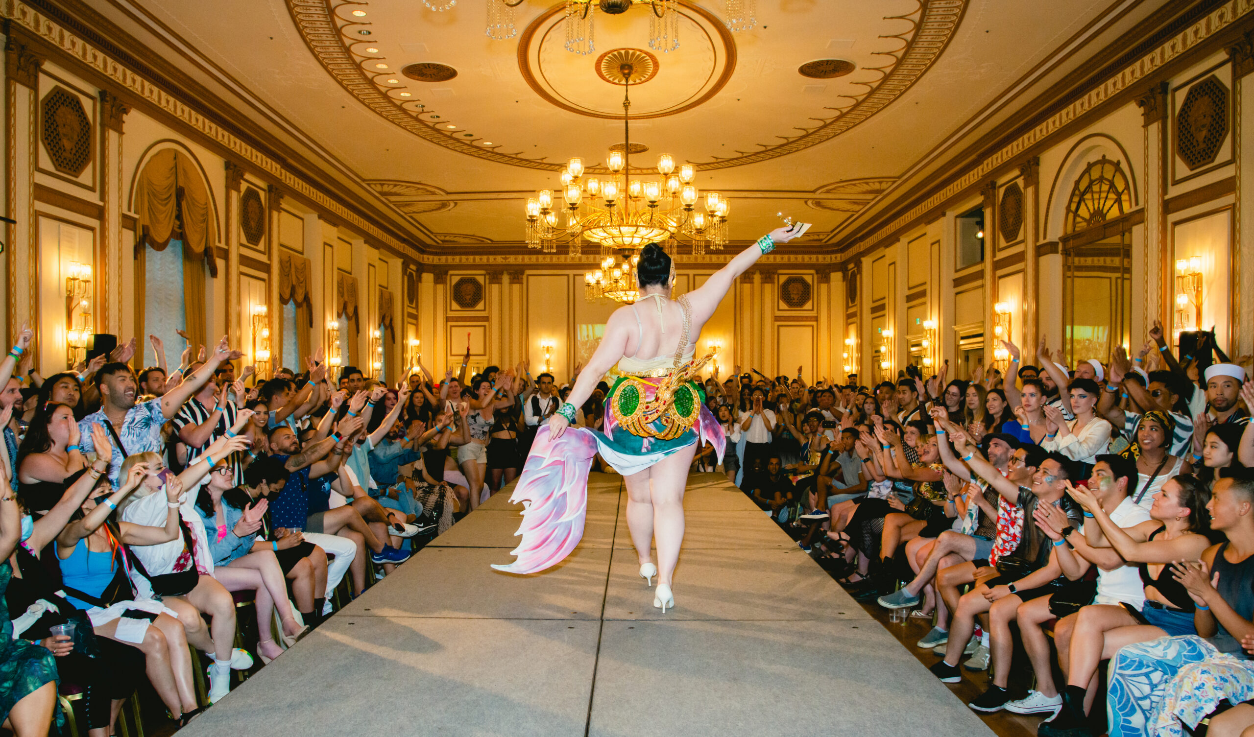 In an opulent ballroom, a performer struts down a runway lined with rows of cheering, grinning audience. Dressed in a colourful short skirt with a hand-painted fish tail, her bare back is visible as she strides away.