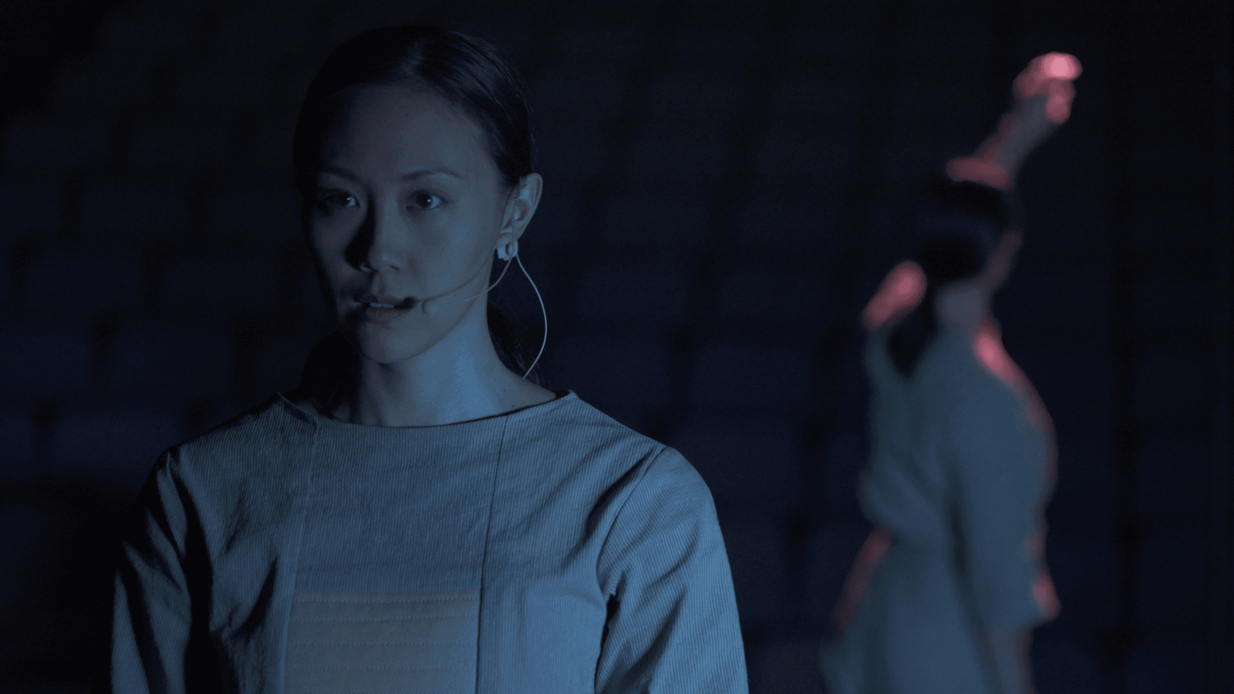 Two asian performers stand on an empty stage under unforgiving light. The first woman wears a severe, focussed expression, underscored by her futuristic structured grey top. In the background, the second performer, dressed in matching garb, faces away and reaches above her head.