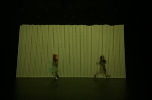 Two blurry figures run in different directions on a dark stage against a striped backdrop like faded yellow wallpaper. The edges of the blurry figures are tinted with red and green light, lending them the quality of a digital glitch.