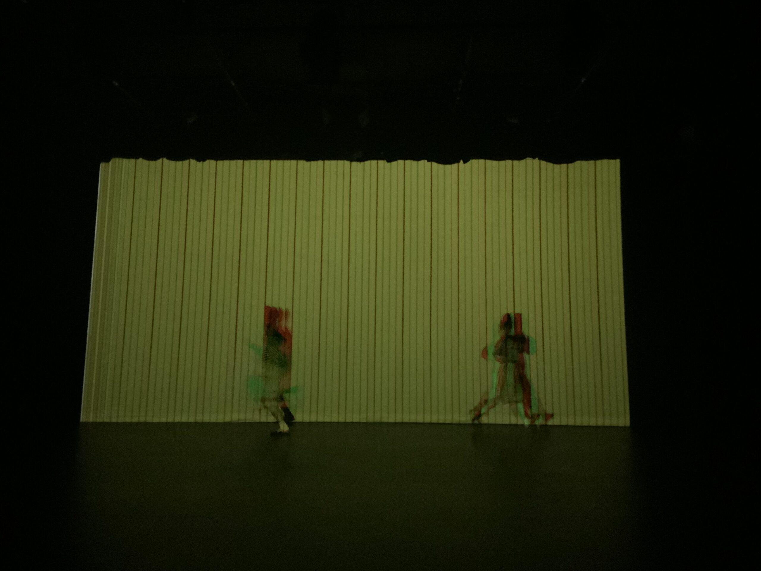 Two blurry figures run in different directions on a dark stage against a striped backdrop like faded yellow wallpaper. The edges of the blurry figures are tinted with red and green light, lending them the quality of a digital glitch.