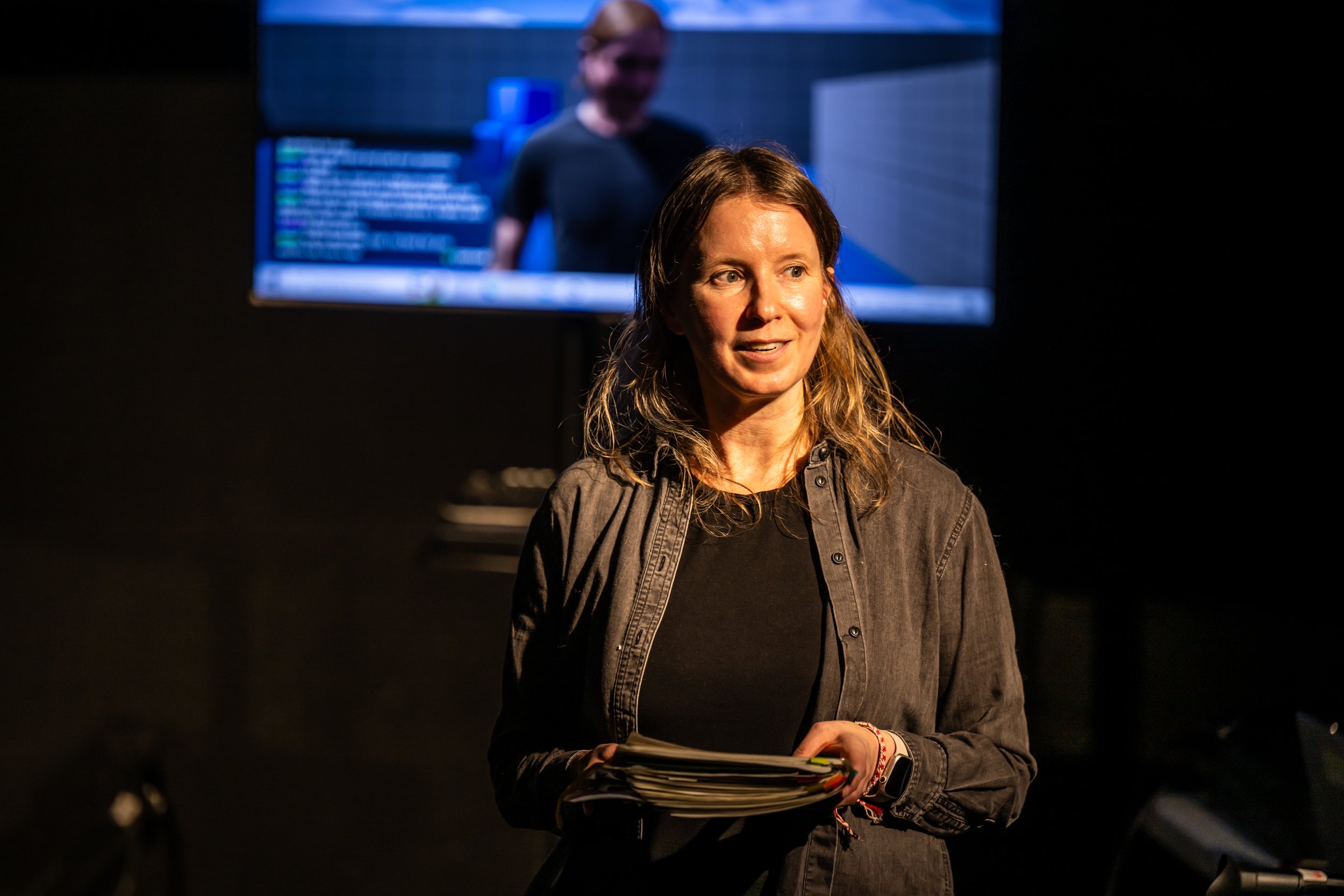 Image description: A woman with a kind expression holds a thick stack of papers as though she's delivering a presentation. Behind her on the stage, an out-of-focus screen shows a plainly-dressed video game character in a sterile environment.