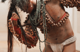 Image description: A solo performer is caught mid-movement, his head turned sideways and body curved. His striking attire leaves much of his tan skin exposed. He wears sleeves adorned with ayoyotes, which are ancestral seeds that rattle, a long beaded turquoise necklace looped many times around his neck, and a white loin cloth. His long hair, worn in a ponytail that streams forward from the top of his head, as well as his skin and attire, are covered with an uneven layer of pale ash.