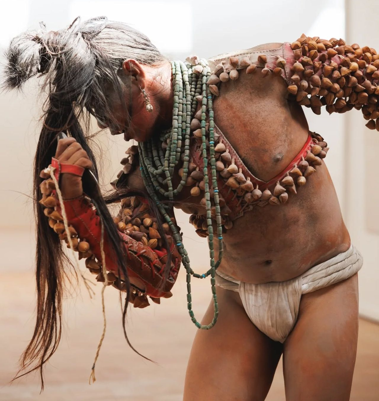 Image description: A solo performer is caught mid-movement, his head turned sideways and body curved. His striking attire leaves much of his tan skin exposed. He wears sleeves adorned with ayoyotes, which are ancestral seeds that rattle, a long beaded turquoise necklace looped many times around his neck, and a white loin cloth. His long hair, worn in a ponytail that streams forward from the top of his head, as well as his skin and attire, are covered with an uneven layer of pale ash.