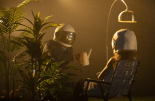 Image description: Two dancers wearing astronaut helmets create an absurd scene: one sits with her back to us in a lawn chair under a reading lamp while the other holds up a watering can, partially obscured by an array of potted tropical plants.