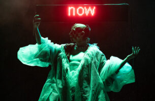 Image description: A trans-masculine drag artist stands on a dark stage, holding an LED-sign where the word "now" glows red. Their bald head is turned in profile, with a checkerboard smile painted across their face. They wear a billowing white coat made of luxuriously gauzy material, which hangs open, exposing a flat, muscular chest.