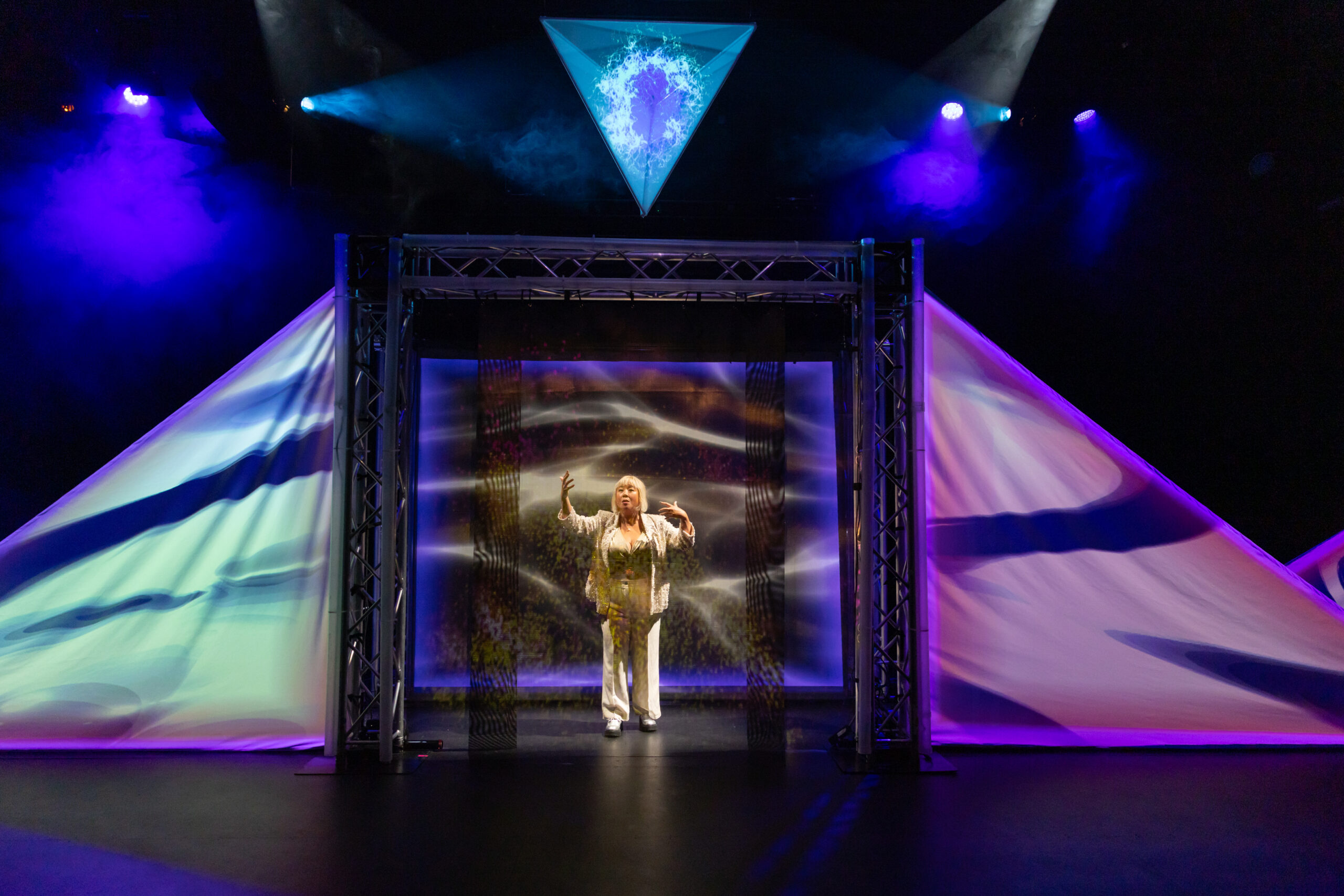 Image description: A woman dressed in a white suit poses playfully with her back towards the audience. She is far back on a stage filled with haze, framed in a wide doorway flanked by triangular white screens, and lit with showy blue and red spotlights.