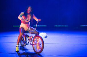 Image description: A Black woman straddles a low-rider bicycle with gold spokes and long shiny handlebars. Dressed in a matching crisp white crop top and short shorts, she looks out to the audience with her mouth open, as though she's in the middle of shouting "hey!".