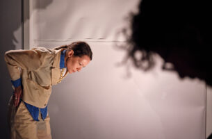 Image description: A light-skinned Black woman dressed impeccably in a tailored tan suit bends at the waist, staring with sharp eyes towards the ground before her, her elbow crooked precisely above her back. In the foreground, a second figure's face, also cast downwards, creates a blurred silouette.