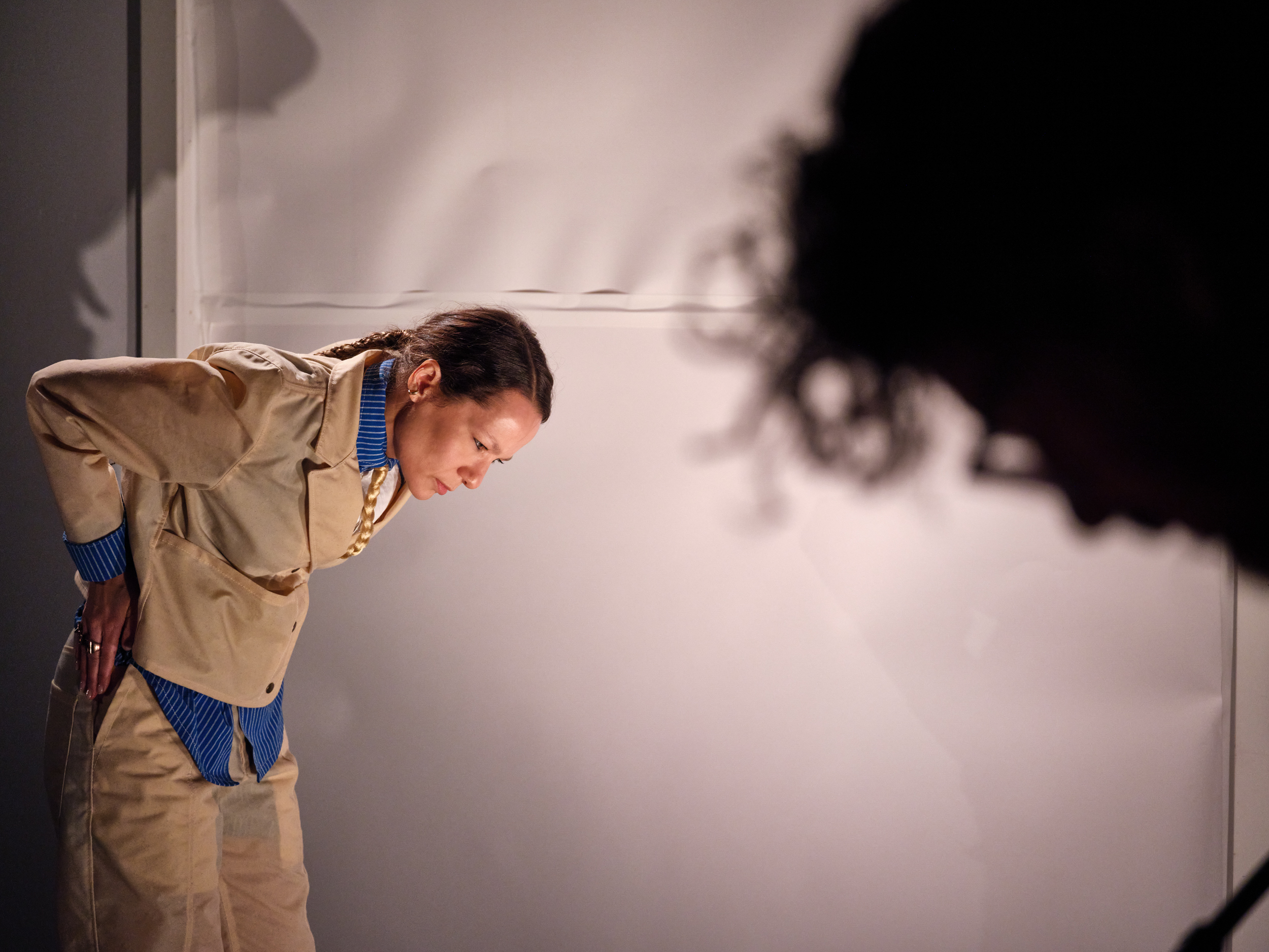 Image description: A light-skinned Black woman dressed impeccably in a tailored tan suit bends at the waist, staring with sharp eyes towards the ground before her, her elbow crooked precisely above her back. In the foreground, a second figure's face, also cast downwards, creates a blurred silouette.