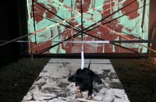Image description: A kneeling performer bows, forehead to ground, in the middle of a stage covered with projected archival photographs of Black people with grave expressions. The floor of the stage is covered in sand, and long strips of tape crisscross the stage above the prostrate performer.