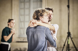 Image description: Two men hold each other like teens at a highschool dance. They pucker their lips, but turn their heads away from eachother, towards the audience. Behind them, a third performer watches anxiously.