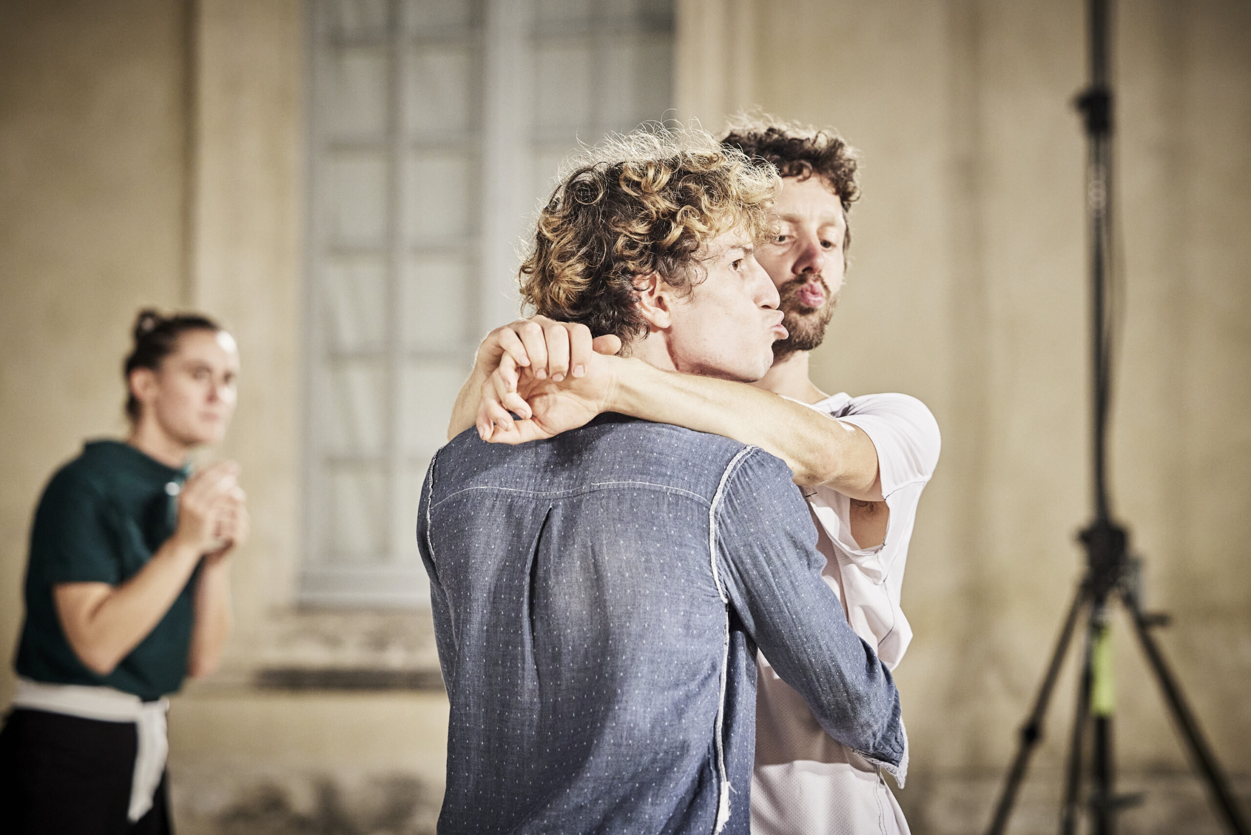 Image description: Two men hold each other like teens at a highschool dance. They pucker their lips, but turn their heads away from eachother, towards the audience. Behind them, a third performer watches anxiously.