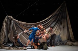 Image description: At the centre of a tangled clump of dancers, a woman wraps her hands around the neck of a man who holds his hands up in a gesture of prayer. Behind them, a huge sheet drapes, dyed with a muted palette of greys and blues.