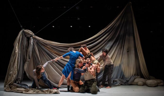 Image description: At the centre of a tangled clump of dancers, a woman wraps her hands around the neck of a man who holds his hands up in a gesture of prayer. Behind them, a huge sheet drapes, dyed with a muted palette of greys and blues.