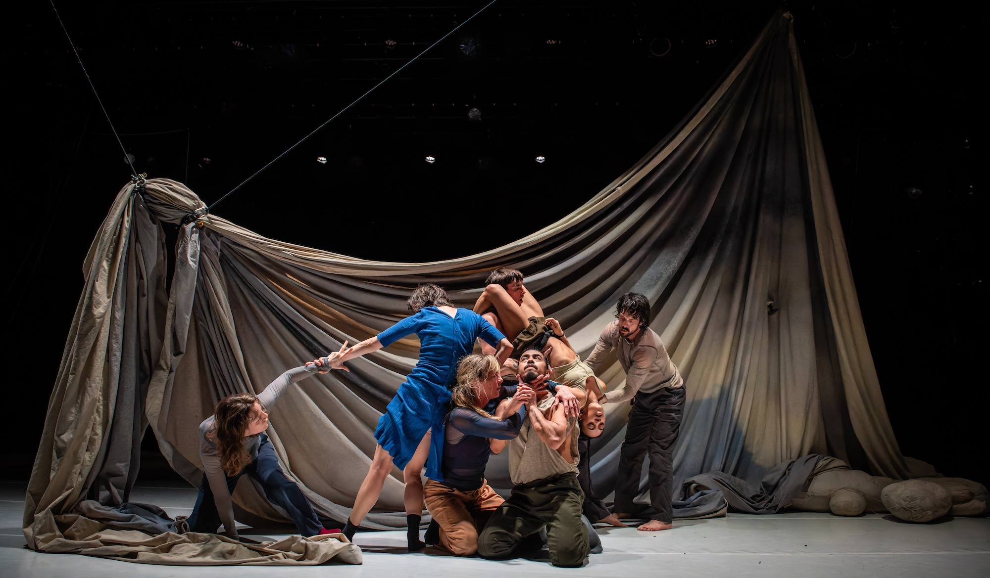 Image description: At the centre of a tangled clump of dancers, a woman wraps her hands around the neck of a man who holds his hands up in a gesture of prayer. Behind them, a huge sheet drapes, dyed with a muted palette of greys and blues.
