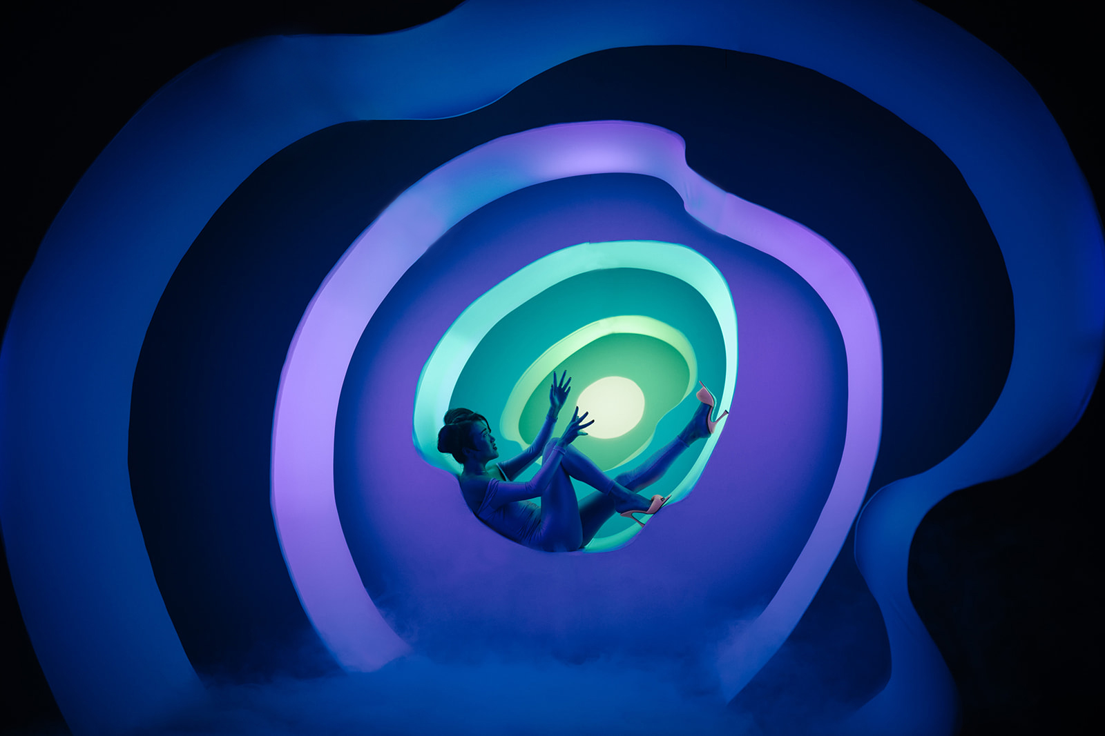 Image description: An Asian woman sits in profile in the centre of a surreal installation: layers of human-sized wavy rings, that get smaller as they receded into the distance, create a portal of hazy blues and greens. Dressed in a flesh-toned leotard and melon-orange high-heels, the woman sits in one of these wavy rings, fitting her spine against the curved bottomw, and stretching her bare leg upward along the curved edge. Her energy appears calm in the centre of this constructed vortex.