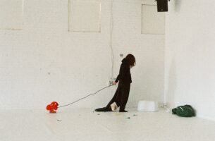 Image description: A performer with dark curly hair wears a leather jacket backwards and a single yellow glove. Their lower half is blocked by the strange things they carry, seemingly chosen as much for their colours and textures more as for their actual purppse: a blue water jug, a bunch of curly gold ribbon, a pink cord, and a crate full of folded clear plastic.