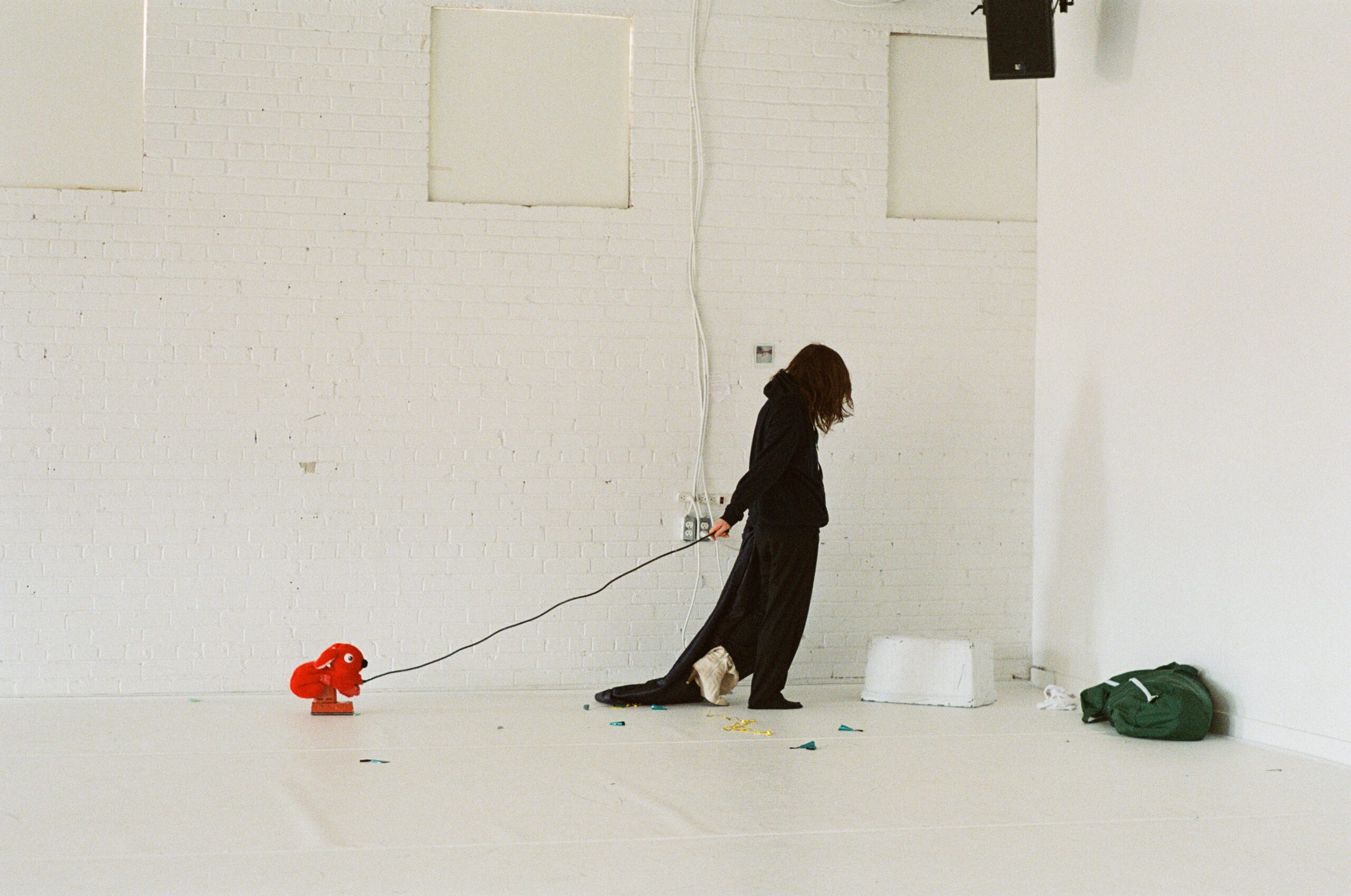 Image description: A performer with dark curly hair wears a leather jacket backwards and a single yellow glove. Their lower half is blocked by the strange things they carry, seemingly chosen as much for their colours and textures more as for their actual purppse: a blue water jug, a bunch of curly gold ribbon, a pink cord, and a crate full of folded clear plastic.