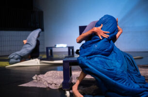 Image description: A performer sits on a blue bench, their face and most of their body covered by a blue sleeping bag. Their hands and one foot, the only parts of them that are visible, are tense, extended wide and folding in towards their shrouded body. Behind them, out of focus, a second dancer sits behind a sleeping bag in a similar position. Around them both, the ground is scattered with large grey sheets.